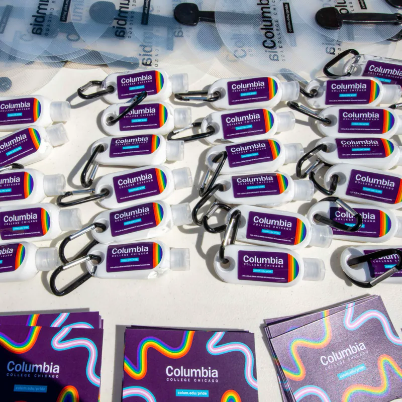 Table of Pride giveaways: rows of Columbia-branded sunscreen bottles on carabiners, clear hand fans, and stacks of rainbow-wave stickers